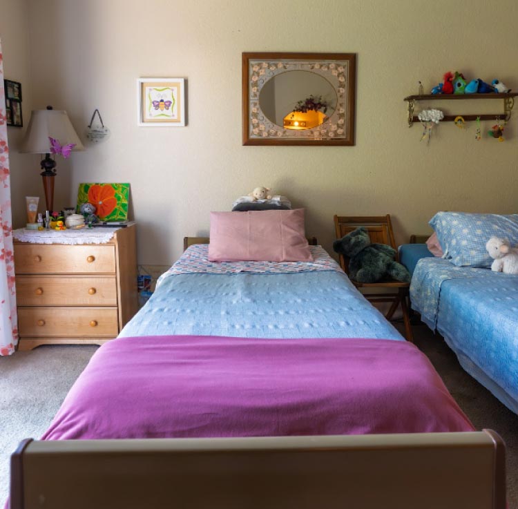 A beautifully staged semi-private resident room featuring two beds with matching violet floral bedding, coordinated window treatments, and a shared dresser, reflecting a clean and welcoming "home-away-from-home" environment.