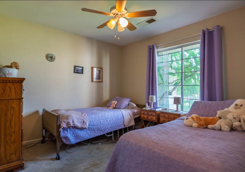 A beautifully staged semi-private resident room featuring two beds with matching violet floral bedding, coordinated window treatments, and a shared dresser, reflecting a clean and welcoming "home-away-from-home" environment.