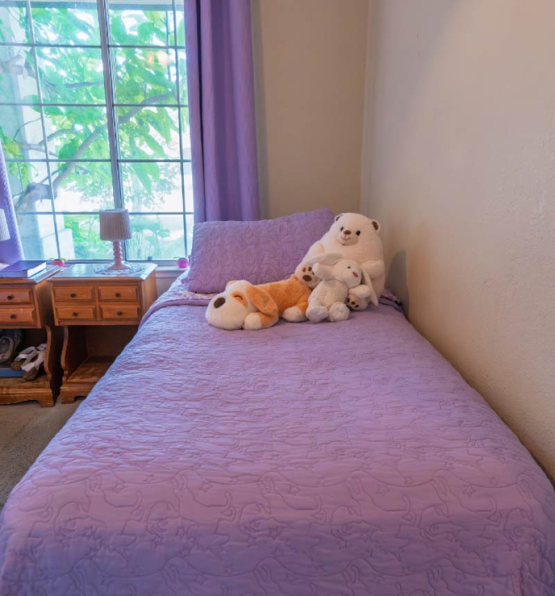 A neat single bed dressed in a quilted purple coverlet and matching pillowcase. Three plush stuffed animals—a cream-colored bear, a white rabbit, and a brown-and-white dog—are arranged near the head of the bed against a plain, neutral wall. Next to the bed, a wooden nightstand sits by a large window with purple curtains, showing green foliage outside