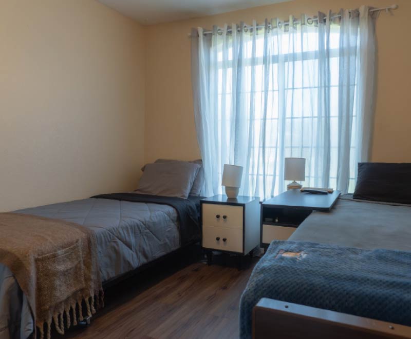 A shared resident bedroom at an assisted living facility featuring two organized twin beds with neutral linens, a central nightstand with lamps, and a large window with sheer white curtains.