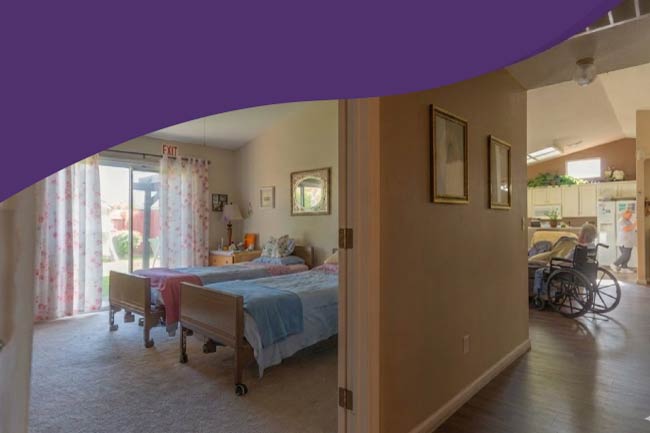 An interior view of a comfortable assisted living room featuring two adjustable medical beds with floral curtains. In the background, a resident in a wheelchair is visible in a hallway leading to a clean, modern kitchen.   