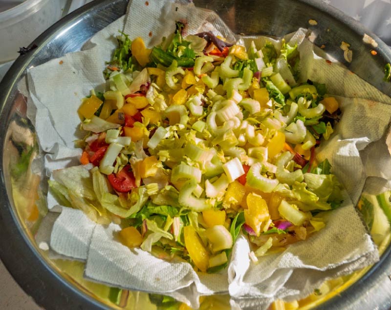 A freshly prepared, nutritious garden salad featuring chopped celery, yellow bell peppers, tomatoes, and leafy greens served in a large metal mixing bowl for senior resident dining.